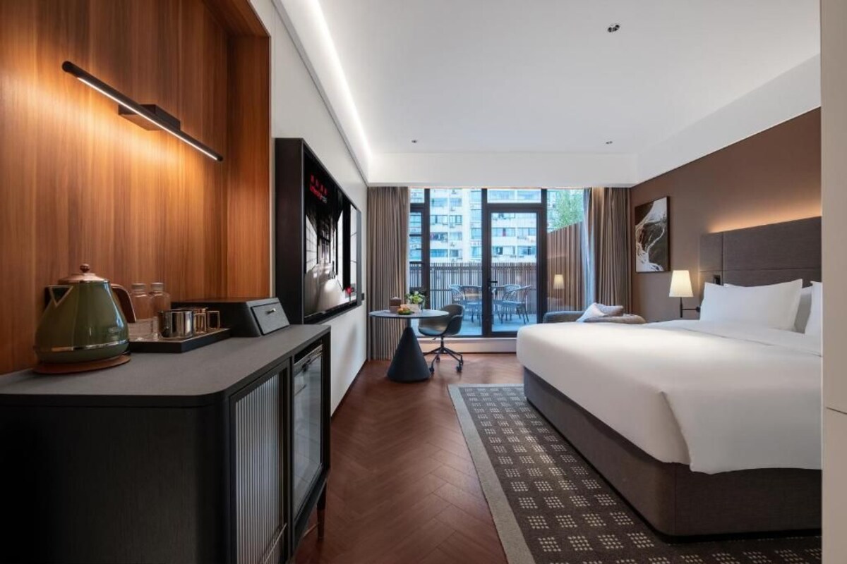 A modern hotel room with a bed, a desk with a kettle, a small table, and a view of the city through large windows.