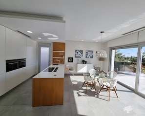 Une cuisine moderne dotée d’un îlot central, de meubles blancs et d’un coin repas avec une table en verre et des chaises blanches.