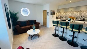 A modern living room with a brown sofa, a round coffee table, and a potted plant. There are four bar stools with green seats and black bases. A kitchen area with patterned backsplash and white cabinets is visible in the background. - Bramble Apartment in Fort William Town Centre (Fort William)