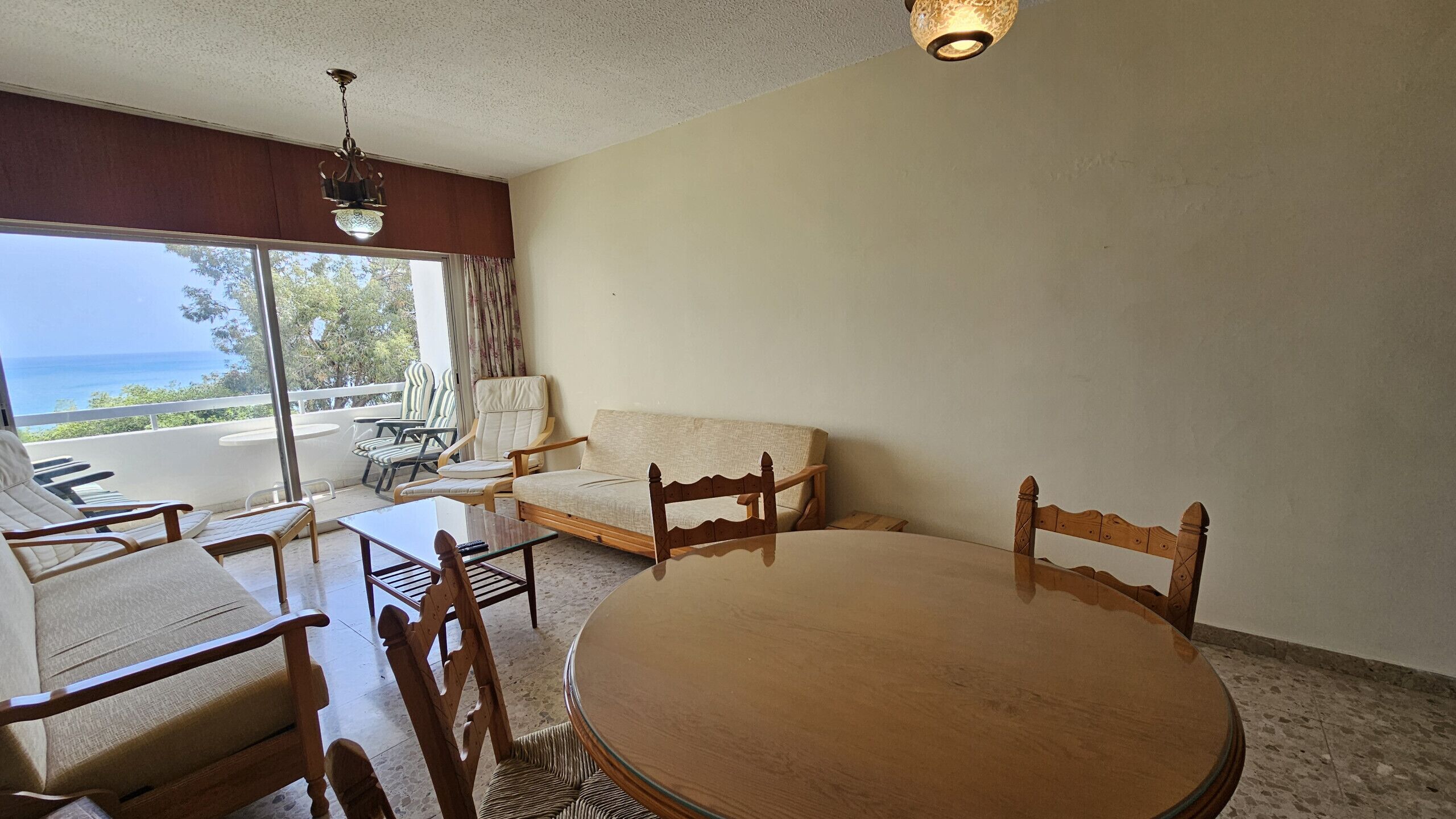 Quarto com mesa redonda, cadeiras de madeira, sofá e varanda com vista para o mar.