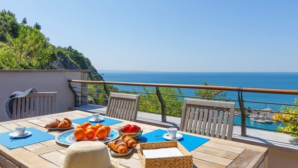 A wooden table with breakfast items, chairs, and a view of the sea.