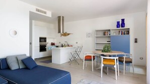 A modern living space with a blue sofa, a dining table with chairs, a kitchen area, and a bookshelf with blue vases.