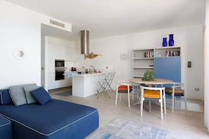 Un espacio habitable moderno con un sofá azul, una mesa de comedor con sillas, una zona de cocina y una estantería con jarrones azules.