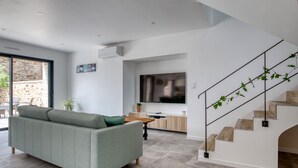 A modern living room with a grey sofa, a wooden coffee table, and a TV mounted on the wall. There is a staircase with a metal railing and a plant on the landing. - Le Logis de la Pierre Bleue - Maison climatisée avec piscine (Férel)