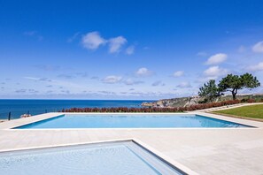 Ein rechteckiger Swimmingpool mit klarem, blauem Wasser, umgeben von einem hellen Fliesenbelag, vor dem sich ein strahlend blauer Himmel und eine ferne Küstenlinie abzeichnen.