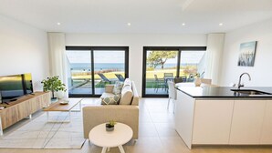 A modern living room with a sofa, coffee table, and a TV. There is a kitchen area with a sink and a view of the outdoors.