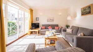 A living room with a sofa, a coffee table, and a TV. There is a window with curtains and a painting on the wall.