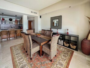 A dining area with a wooden table, wicker chairs, a patterned rug, and a kitchen in the background. - The Beachfront Oasis Ocean Views & Pool Access (Puerto Peñasco)