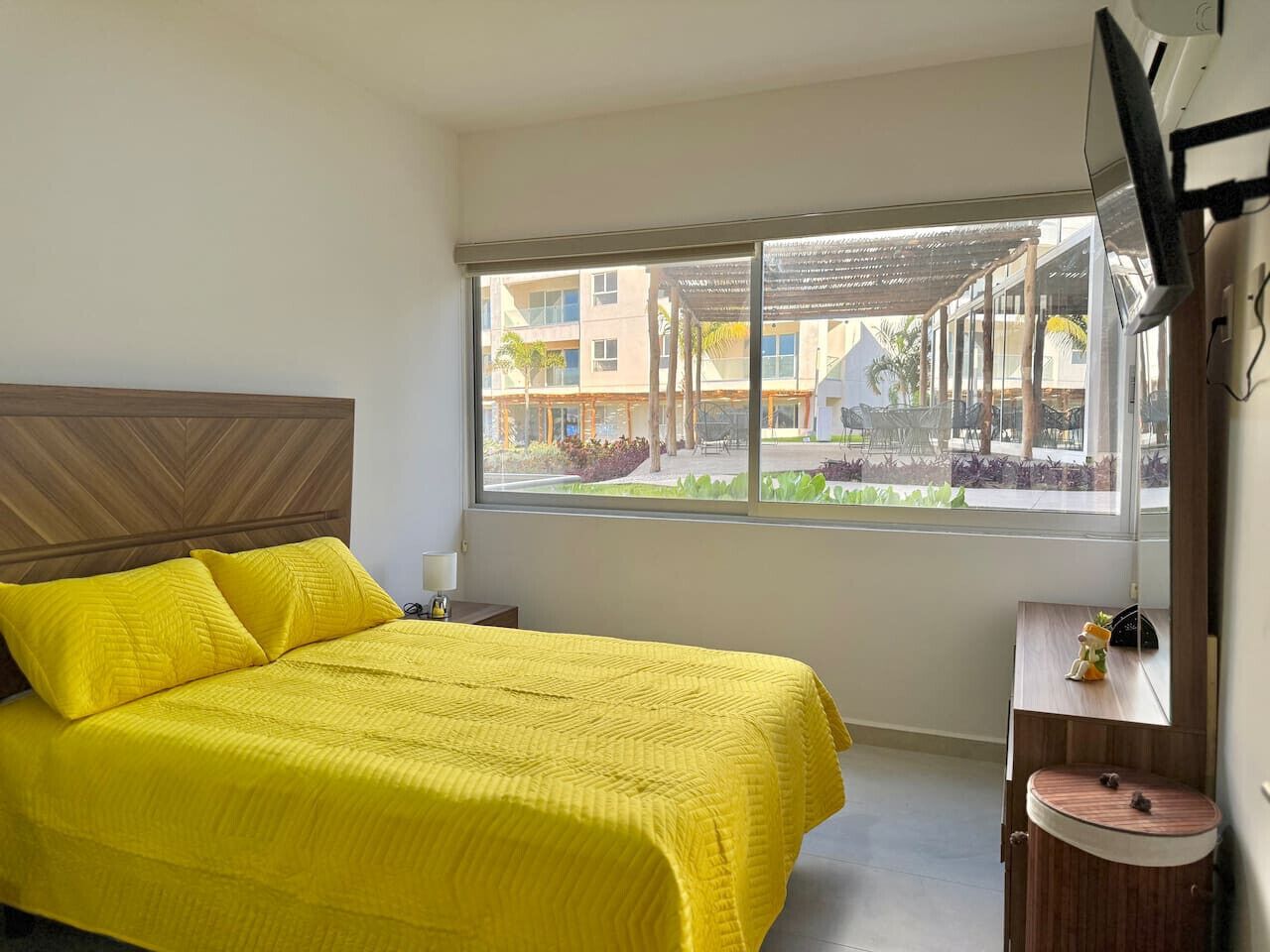 A hotel room with a large bed, a small bedside table, a window with a view of the outdoor area, and a wall-mounted air conditioning unit.