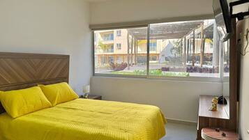 A hotel room with a large bed, a small bedside table, a window with a view of the outdoor area, and a wall-mounted air conditioning unit.