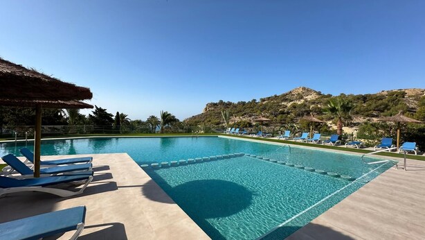 Una piscina con sillas reclinables azules y una sombrilla de paja, rodeada de follaje y colinas.