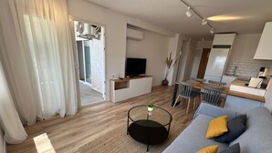 Una sala de estar moderna con un sofá, una mesa de centro y un televisor. Hay un comedor con una mesa y sillas, y una cocina con un refrigerador y un lavabo.