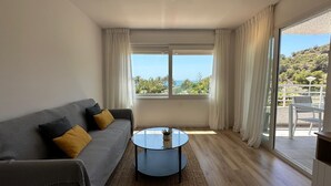 Una sala de estar moderna con un sofá gris, una mesa de centro redonda y un amplio ventanal con vistas a vegetación y una colina.