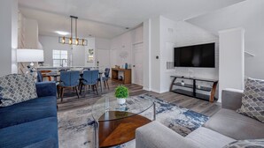 A modern living room with a sofa, coffee table, and TV. There is a dining area with chairs and a table.