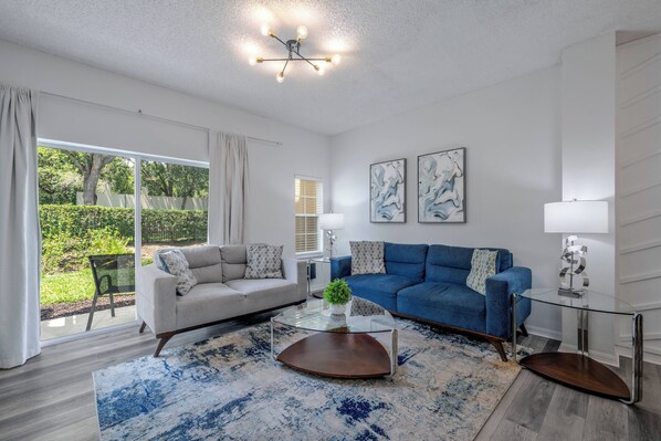 A modern living room with a glass coffee table, a blue sofa, and a beige sofa. There is a large window with curtains, a glass door leading to a garden, and a ceiling light fixture.