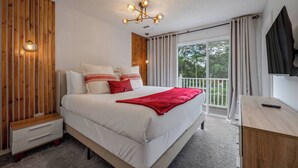 A bedroom with a bed, a wooden bedside table, a desk, and a large window with curtains.