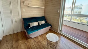 Una habitación con piso de madera, un sofá turquesa con almohadones, una pequeña mesa redonda y un ventanal corredero a un balcón con vista a edificios.