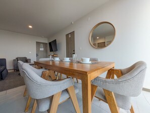 Un comedor con una mesa de madera y sillas tapizadas en gris, un espejo redondo en la pared y un televisor al fondo.