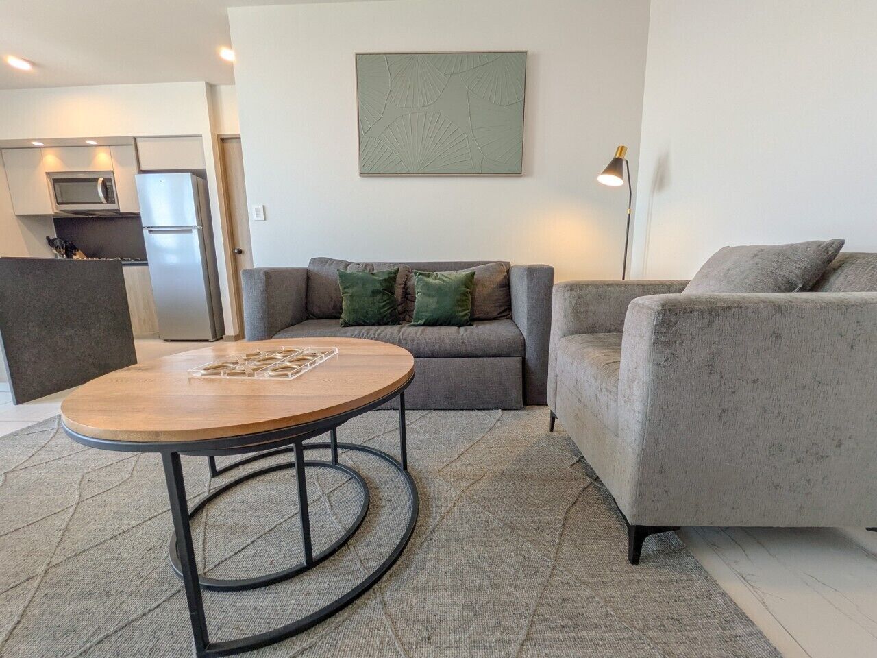A modern living room with a round wooden coffee table, grey sofas, and a kitchen in the background.