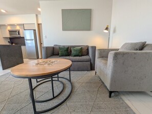 A modern living room with a round wooden coffee table, grey sofas, and a kitchen in the background.