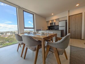 A dining area with a table and chairs, a kitchen with appliances, and a view of the city. - Beautiful Luxury Apartment Near Andares! (Zapopan)