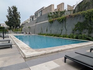 Ein rechteckiger Swimmingpool mit Fliesenrand, umgeben von Liegestühlen und einer Betonmauer mit Bepflanzung.