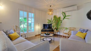 Ein Wohnzimmer mit Sofa, Sesseln, Couchtisch, Fernseher und einem großen Fenster mit Blick auf Grünflächen.