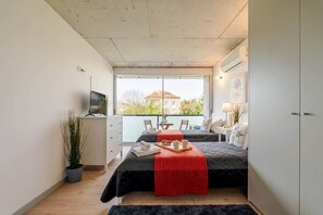 Un dormitorio con dos camas, un televisor, un escritorio, una silla, una planta y un ventanal con vistas.