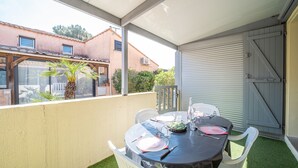 An outdoor patio with a round table set for dining, surrounded by white chairs, and a covered area with a view of neighboring buildings. - Notre Patio - Maisonette climatisée proche plage (Saint-Cyprien)