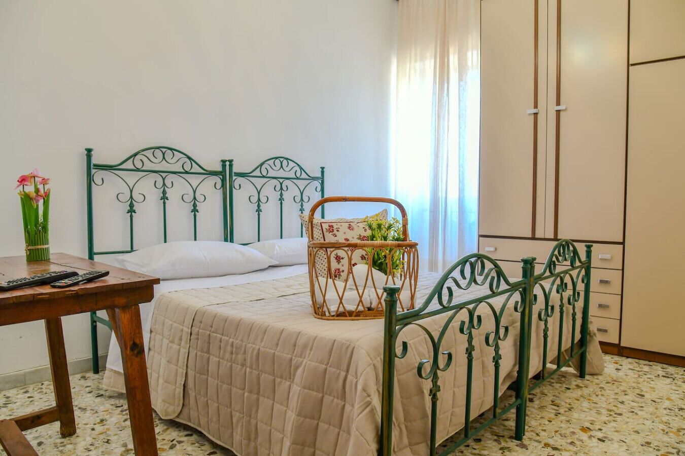 Une chambre avec un lit, une table de chevet ornée d’un vase, un panier en osier et une armoire.