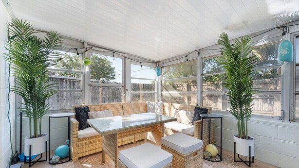 A covered patio area with wicker furniture, including a sofa and a table, surrounded by large windows and potted plants.