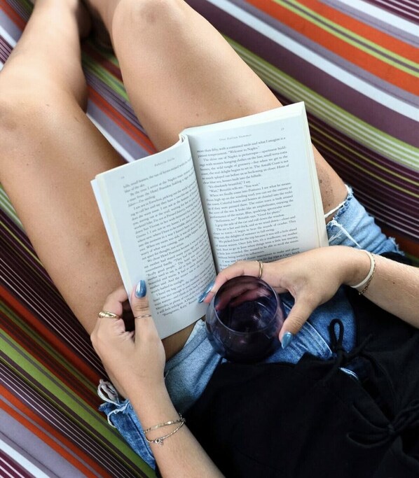 Een persoon ligt in een gestreepte hangmat, leest een boek en houdt een glas vast.