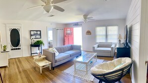 A living room with a grey sofa, a white coffee table, a white armchair, and a black rocking chair. There is a door with a white frame and a small table with a plant.