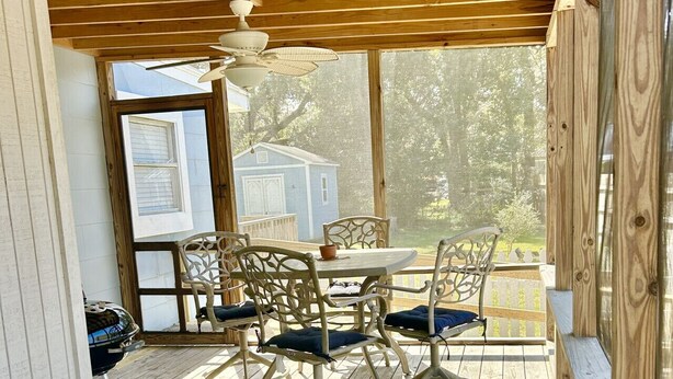 A covered outdoor patio with a wooden ceiling, a table with chairs, and a view of a blue house in the background.