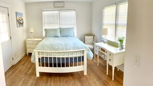 A bedroom with a white bed, a desk, and a chair. There are two windows with blinds, a ceiling fan, and a wall-mounted light.