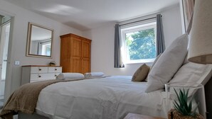 Ein Schlafzimmer mit einem Bett, einem Holzschrank, einem Fenster mit Vorhängen, einem Nachttisch mit Lampe und einem Spiegel an der Wand.