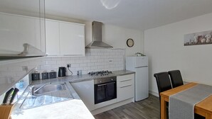 Eine kompakte Küche mit weißen Schränken, einem Gasherd, einem Backofen und einem Kühlschrank. Ein Esstisch mit zwei Stühlen und eine Uhr sind an der Wand angebracht.
