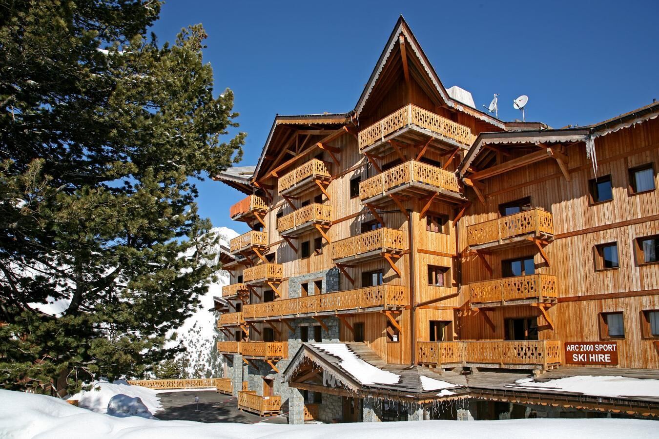 A multi-story wooden building with balconies, a ski hire sign, and snow-covered surroundings.