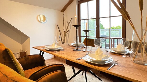 A dining area with a wooden table set for four, chairs, a clock on the wall, and a window with natural light.