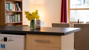 Eine Küchenzeile mit einer Arbeitsplatte, einer Vase mit gelben Blumen, einer Packung Walker's Finger Biscuits und einem Bücherregal im Hintergrund.