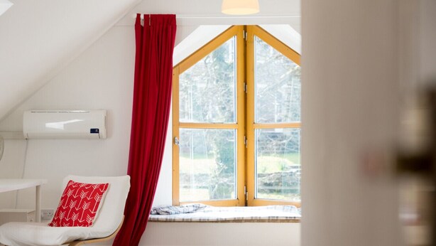 Ein Zimmer mit einem weißen Sessel, einem roten Vorhang und einem großen Fenster mit Blick auf Bäume.