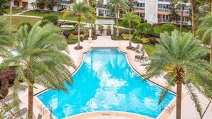 A large swimming pool surrounded by palm trees and lounge chairs, with a building complex in the background. - Condo with Golf Course Views, Near Attractions (Kissimmee)