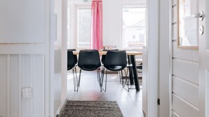 Una habitación con una puerta blanca, una ventana con cortina rosa, una mesa de madera y cuatro sillas negras.