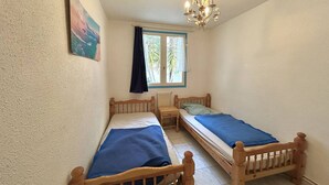 Une petite pièce accueillante, avec deux lits en bois, une petite table, une chaise, une fenêtre avec des rideaux et un luminaire au plafond.