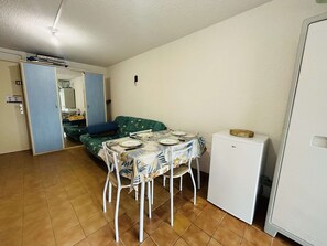 A compact living space with a dining table set for two, a sofa, a small refrigerator, and a door leading to another room.