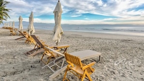 A beach with rows of wooden lounge chairs and white umbrellas, a sandy shore, and a calm sea under a partly cloudy sky. - 3BR beach condo - pool, shore access & bay views (Cruz de Huanacaxtle)