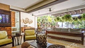 A modern living room with a sofa, chairs, and a coffee table. There is a television on the left and a large window with plants. - 3BR beach condo - pool, shore access & bay views (Cruz de Huanacaxtle)