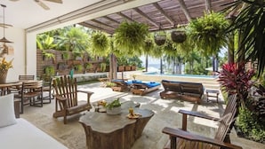 An outdoor patio area with wooden furniture, hanging plants, and a view of a pool. - 3BR beach condo - pool, shore access & bay views (Cruz de Huanacaxtle)