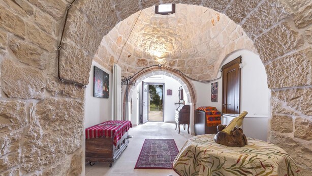 A room with stone walls, a wooden bench, a bed with a patterned bedspread, a red and white checkered tablecloth, a small rug, and a doorway leading to another room.
