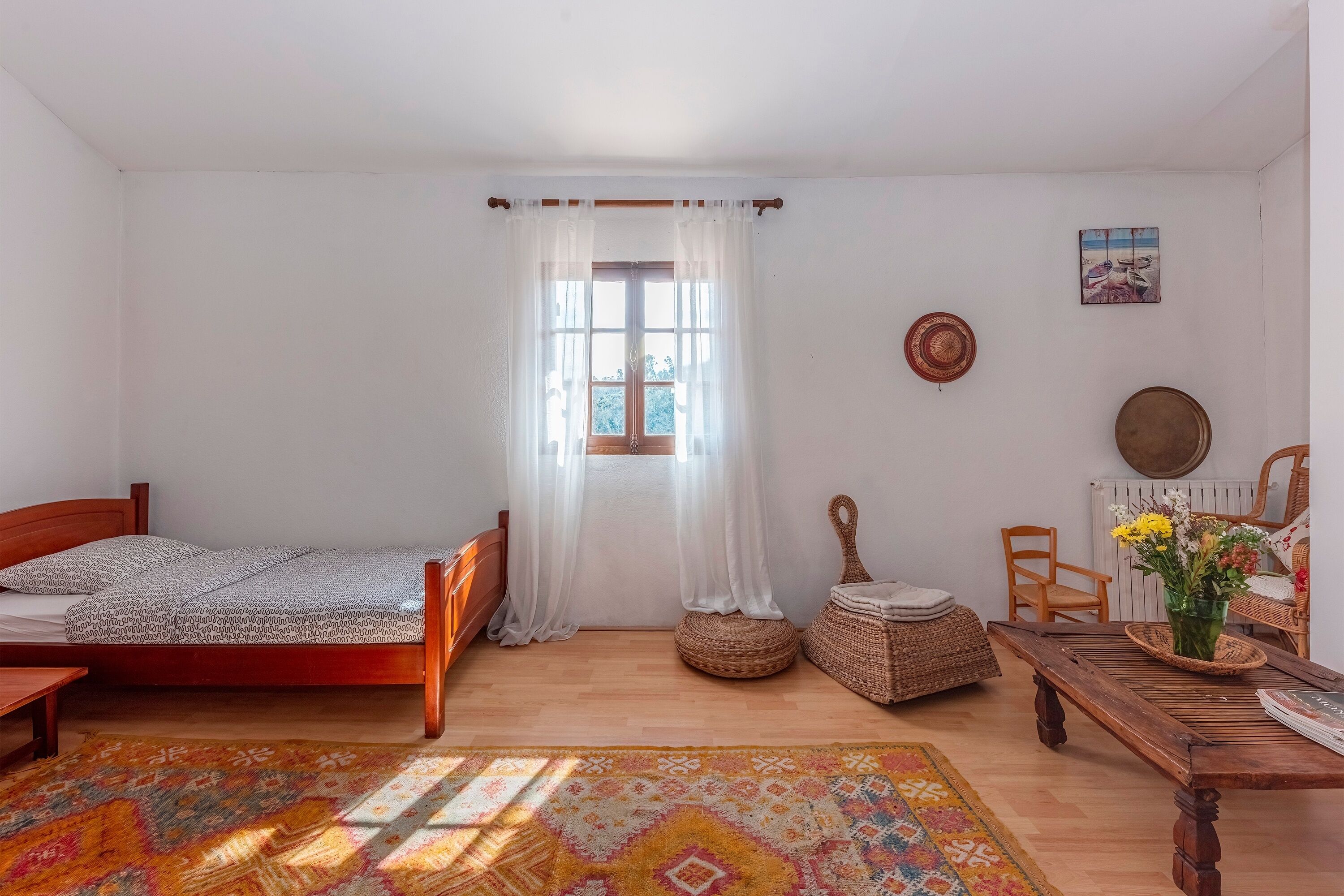 A bedroom with a wooden bed, a window with sheer curtains, a woven basket, a wooden table with a vase of flowers, and a patterned rug.